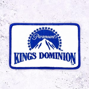 Pode incluir: Remendo retangular com fundo branco e borda azul. O remendo apresenta o texto "Paramount's KINGS DOMINION" em azul, com um gráfico de montanha e um design de estrela acima do texto.