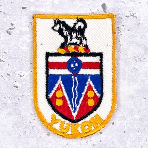 Könnte beinhalten: Gestickter Aufnäher mit dem Wappen von Yukon. Das Design umfasst einen schwarzen Hund, rote und blaue Streifen sowie das Wort "YUKON" in Gelb. Der Aufnäher hat einen weißen Hintergrund und einen gelben Rand.