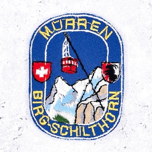 Op de afbeelding: Geborduurde patch met een blauwe achtergrond met de woorden "Murren" en "Birg-Schilthorn" in geel. De patch toont een rode kabelbaan, besneeuwde bergen en twee schilden met witte kruisen en een zwarte beer.