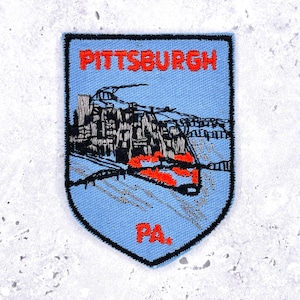 Könnte beinhalten: Gestickter Aufnäher mit hellblauem Hintergrund und schwarzem Rand. Der Aufnäher zeigt den Städtenamen "PITTSBURGH" in Rot, zusammen mit einer schwarz-grauen Illustration der Skyline und Brücken der Stadt. Die Buchstaben "PA" sind rot.