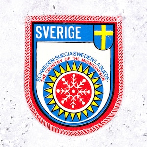 Peut inclure: Un écusson brodé en forme de bouclier avec le mot SVERIGE en blanc, un emblème de croix bleue et jaune, et un cercle rouge central avec un flocon de neige blanc entouré d'un soleil jaune, portant l'inscription The Country of the Midnight Sun.