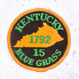 May include: A round patch with an orange border and a black center. The patch features the state of Kentucky in orange, with the text "KENTUCKY" above and "BLUE GRASS" below in green. The year "1792" and the number "15" are also in green.