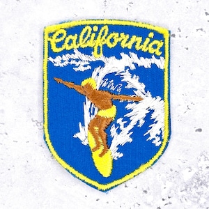 May include: A shield-shaped embroidered patch with a blue background and a yellow border. It features a brown surfer on a yellow surfboard riding a white wave, with the yellow cursive text California embroidered across the top section.