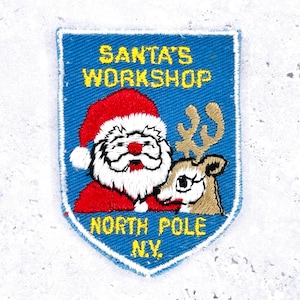 Pode incluir: Patch bordado com o Pai Natal e uma rena, com o texto "SANTA'S WORKSHOP NORTH POLE N.Y." em amarelo sobre fundo azul. O Pai Natal usa um chapéu vermelho e tem barba branca. A rena é castanha.