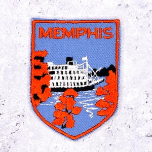 May include: Embroidered patch featuring a steamboat on a light blue background, with the word "MEMPHIS" in orange. The patch has an orange border and orange floral accents. A souvenir item.