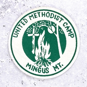 Può includere: Una toppa circolare con un design verde e bianco. Il testo "UNITED METHODIST CAMP" e "MINGUS MT." circonda un'immagine centrale di una croce, un albero e un falò. La toppa è probabilmente per un campo o un'organizzazione.