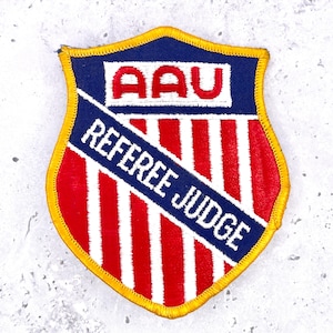 May include: A shield-shaped embroidered patch with a yellow border. The top section is blue with the red text AAU. The lower section features red and white vertical stripes overlaid by a diagonal blue banner with the white text REFEREE JUDGE.