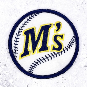 May include: A circular embroidered patch featuring a white baseball design with dark blue stitching. The center displays the text M's in bold dark blue lettering with a bright yellow outline, all enclosed by a thick dark blue stitched border.