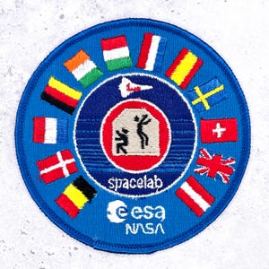 May include: A circular blue patch featuring the word "spacelab" and the logos of ESA and NASA. The patch includes flags of various countries around a central emblem with two figures. The patch is embroidered.