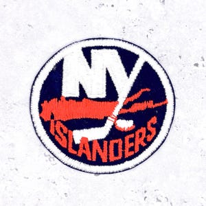 Może przedstawiać: Okrągła naszywka z logo New York Islanders. Logo zawiera białe litery "NY", pomarańczową kij hokejowy i pomarańczowy napis "ISLANDERS". Naszywka jest obramowana granatowym okręgiem z białym obrysem.
