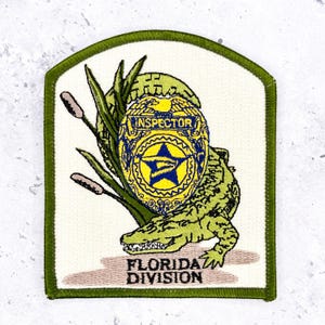 May include: An embroidered patch with a green border on a white background featuring a green alligator, tall green reeds, and a yellow and blue badge with the text INSPECTOR. Below the alligator, the text FLORIDA DIVISION is embroidered in black.