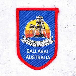 May include: A blue and red patch featuring a gold mining tower over a sunset design. The patch reads "SOVEREIGN HILL BALLARAT AUSTRALIA" in white text. The patch has a red border and a shield shape.