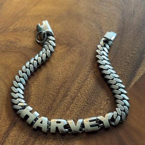 May include: Silver-toned chain bracelet with the name "HARVEY" spelled out in bold, block letters. The bracelet features a curb link design and a secure clasp. The bracelet is displayed on a wooden surface.