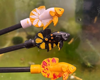 May include: Three colourful betta fish ornaments. One is white and yellow, another black and yellow, and the third is orange with red detailing. Each fish is attached to a black rod, possibly for display or use, against a blurred green background.
