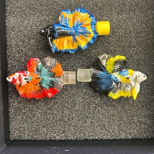 May include: Three colorful betta fish figurines with intricate details. One is black and blue with yellow fins, another is orange, red, and white, and the third is blue and yellow. Each has a clear, cylindrical connector.