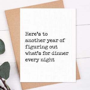 May include: A white greeting card with the text "Here's to another year of figuring out what's for dinner every night" is placed on a brown envelope. A sprig of green leaves is in the lower left corner. The background is a white wooden surface.