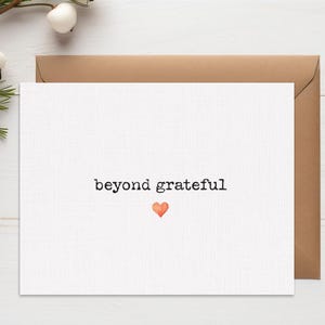 May include: A white greeting card with the words "beyond grateful" in black text and a small orange heart. The card is placed on a brown envelope, with decorative pinecones and greenery in the background.