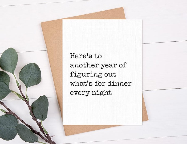 May include: A white greeting card with the text "Here's to another year of figuring out what's for dinner every night" is placed on a brown envelope. A sprig of green leaves is in the lower left corner. The background is a white wooden surface.