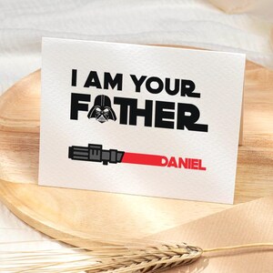May include: A white greeting card with the text "I AM YOUR FATHER" in black, with a Darth Vader graphic. Below, a red lightsaber graphic and the name "DANIEL" in red. The card is on a wooden plate.