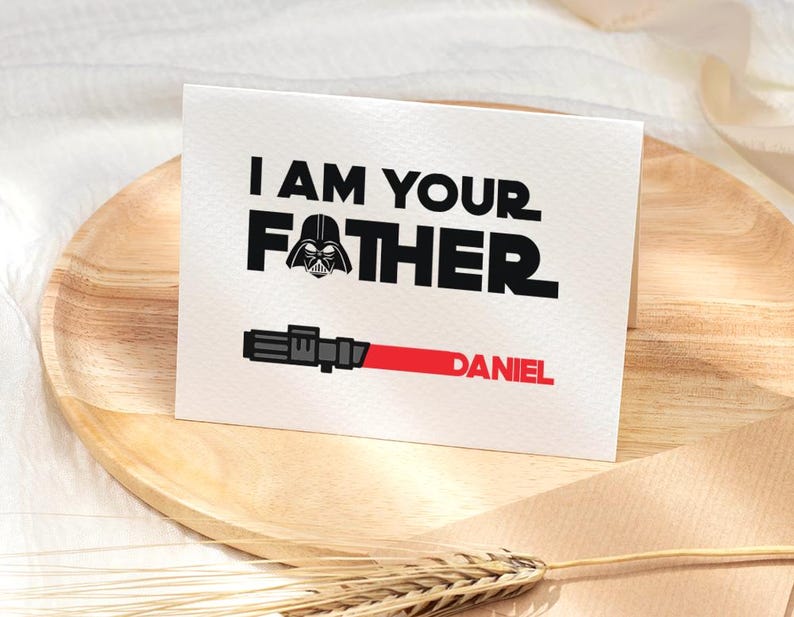 May include: A white greeting card with the text "I AM YOUR FATHER" in black, with a Darth Vader graphic. Below, a red lightsaber graphic and the name "DANIEL" in red. The card is on a wooden plate.