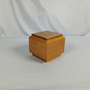 May include: A square wooden box with a natural light brown grain and a smooth finish. The box features a tiered base design and a flat, square lid that sits flush on top. The wood displays a consistent warm tone with visible linear grain patterns.