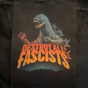 May include: A black patch featuring a Godzilla-like monster in blue and orange, with the text "DESTROY ALL FASCISTS" in orange and yellow. The patch is rectangular and appears to be made of fabric.