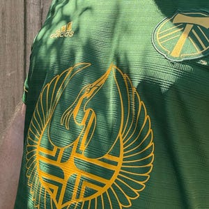 May include: Green soccer jersey with a gold and green logo of a bird and the number 7. The Adidas logo is visible on the upper left chest. The jersey has a textured pattern.
