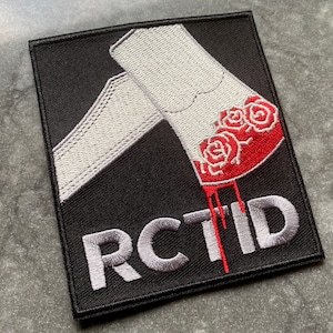 May include: Embroidered patch with a black background and a white and red design. The design features a stylized image of a white object with red roses and the letters "RCTID" in gray.