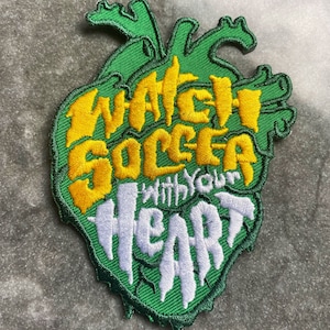 Op de afbeelding: Geborduurde patch in de vorm van een groen hart met de woorden "Watch Soccer With Your Heart" in geel en wit. De patch heeft een gedetailleerd, gestructureerd ontwerp.