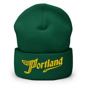 Puede incluir: Un gorro verde oscuro con un borde doblado. La palabra "Portland" está bordada en escritura amarilla en el borde. El gorro está hecho de un material suave y tejido.
