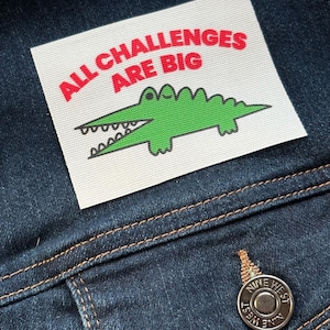 Puede incluir: Un parche rectangular blanco con el texto "ALL CHALLENGES ARE BIG" en letras rojas y un cocodrilo verde de dibujos animados. El parche está cosido a una chaqueta vaquera azul oscuro.