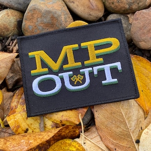 May include: A black rectangular patch with the words "MP OUT" embroidered in yellow, green, and white. The patch features a small emblem of crossed axes. The patch is surrounded by autumn leaves and rocks.