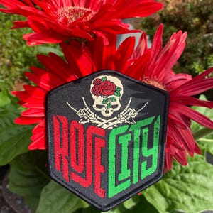Op de afbeelding: Een zwarte, zeshoekige patch met een schedel met roos en gekruiste botten. De patch heeft de tekst "ROFF CITY" in rode en groene blokletters. De patch staat tegen een achtergrond van levendige rode bloemen en groen gebladerte.