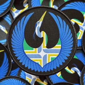 May include: Round black patch featuring a blue heron design with outstretched wings. The heron has a yellow beak and is set against a background of blue, green, and yellow geometric shapes. The patch is likely an embroidered emblem.
