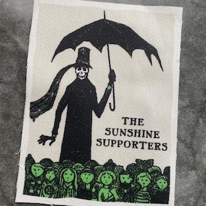 Op de afbeelding: Rechthoekige patch met een zwarte illustratie van een skeletfiguur met een paraplu. De figuur draagt een hoge hoed en een lange sjaal. Daaronder een menigte groen omrande figuren. Zwarte tekst: "THE SUNSHINE SUPPORTERS".