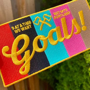 Op de afbeelding: Rechthoekige patch met een gele rand en het woord "Goals!" in gele letters. De patch heeft een kleurblokontwerp met rode, zwarte, turquoise, roze en bruine strepen. Tekst bevat "5 at a time we want" en "Est. 1975 Portland."