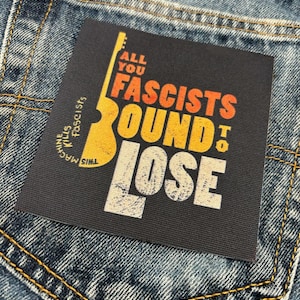 May include: A dark grey patch with the words "ALL YOU FASCISTS BOUND TO LOSE" in orange and white, and a yellow guitar graphic. The patch is on a denim jeans pocket.