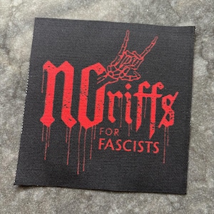 May include: Black square patch with red text and a skeleton hand gesture. The text reads "no riffs for fascists" in a gothic font with dripping blood effect. The patch is on a gray surface.