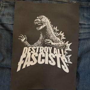 May include: Black rectangular patch featuring a white illustration of Godzilla with the text "DESTROY ALL FASCISTS". The patch is placed on a denim jacket.
