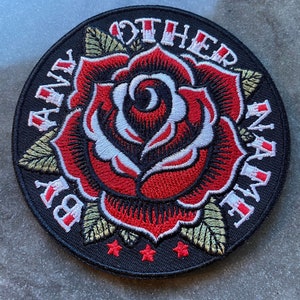 May include: Round embroidered patch featuring a red rose with white and black details. The patch has the text "BY ANY OTHER NAME" in a circular pattern around the rose. The patch has a black background with three red stars.