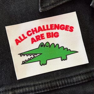 May include: A rectangular white fabric patch featuring the red text ALL CHALLENGES ARE BIG above a green cartoon crocodile illustration with black outlines. The patch is attached to a black denim garment with visible stitching and a collar.