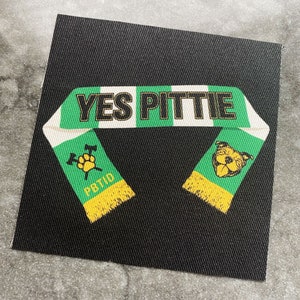 Op de afbeelding: Een zwarte vierkante patch met een groen en wit sjaalontwerp. De tekst "YES PITTIE" staat op de sjaal. De sjaal heeft gele franjes en groene secties met een pootafdruk en een hondengezicht.