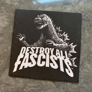 Pode incluir: Patch quadrado preto com uma ilustração branca de um monstro semelhante a Godzilla. O texto "DESTROY ALL FASCISTS" é impresso em letras brancas em negrito. O patch é provavelmente feito de tecido.