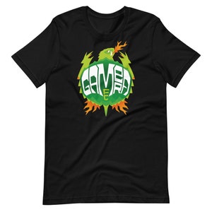 Puede incluir: Camiseta negra con un diseño gráfico verde y naranja. El diseño incluye la palabra "GAMERA" en blanco, rodeada por una criatura verde con llamas naranjas. La camiseta está hecha de un material suave.