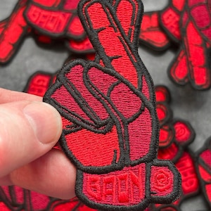 Op de afbeelding: Rode geborduurde patch in de vorm van een hand die het vredesteken maakt. De patch heeft een zwarte rand en het woord "GEON" is geborduurd aan de basis van de hand. De patch wordt in een hand gehouden.