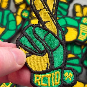 May include: Embroidered patch featuring a hand gesture with the index and middle fingers raised, forming a peace sign. The fingers are yellow, outlined in black, and set against a green background. The patch includes the text "RCTID" in yellow.