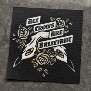 May include: Black square patch with the text "All Crows Are Brilliant" in a stylized font. The design includes a crow skull and floral elements in cream and tan. The patch has a textured surface.