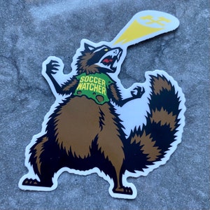 Op de afbeelding: Een sticker met een cartoon wasbeer met een bruin lichaam, een zwart-bruin gestreepte staart en een groen vest met de tekst "SOCCER WATCHER". De wasbeer schreeuwt en heeft een gele straal die uit zijn mond komt.