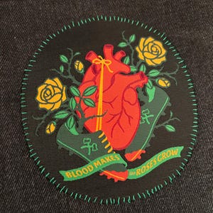 Op de afbeelding: Een ronde patch met een rood hart, gele rozen en groene bladeren. De patch heeft de tekst "BLOOD MAKES the ROSES GROW" op een groene banner. De patch is gestikt met groene draad op een zwarte achtergrond.