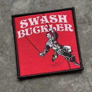 Op de afbeelding: Een rood geborduurd embleem met de witte woorden "SWASH BUCKLER", met een cartoonillustratie van een persoon die aan touwen zwaait. De patch heeft een zwarte rand.